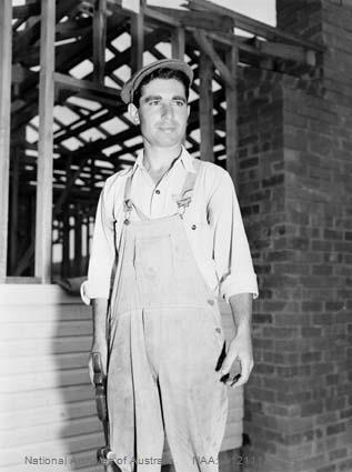 Black and white photograph of Carmel Sciberras, a Maltese carpenter in work overalls, standing on a housing construction site in Canberra