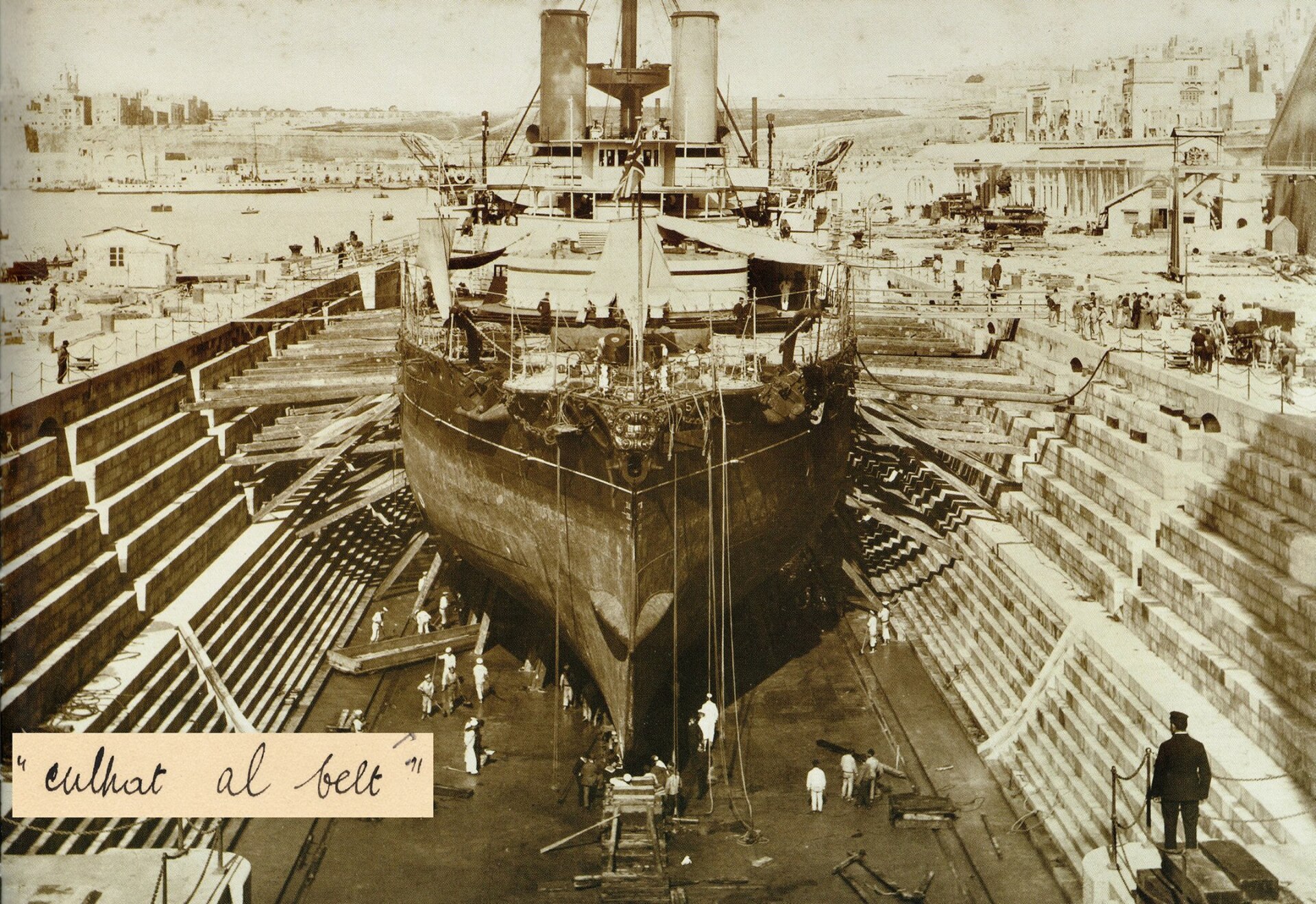A warship in the naval dockyard of Malta, with the harbour fortifications behind.